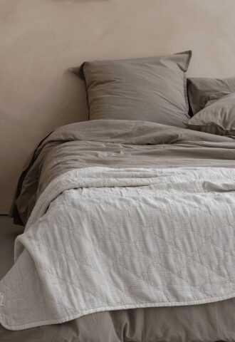 Lit avec draps beiges et couverture matelassée blanche.