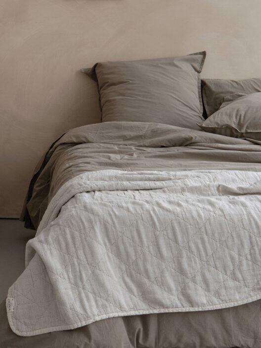 Lit avec draps beiges et couverture matelassée blanche.